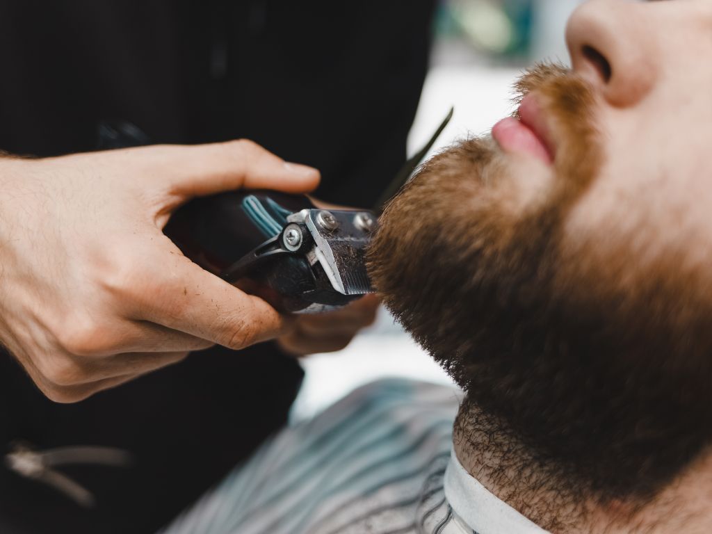 Primo piano di una persona che riceve una rifinitura della barba da Barbiere a Fiumefreddo di Sicilia. Una mano maneggia con maestria un tagliacapelli elettrico, scolpendo la folta barba di un uomo vestito con un classico mantello da barbiere, sullo sfondo di dettagli leggermente sfocati.