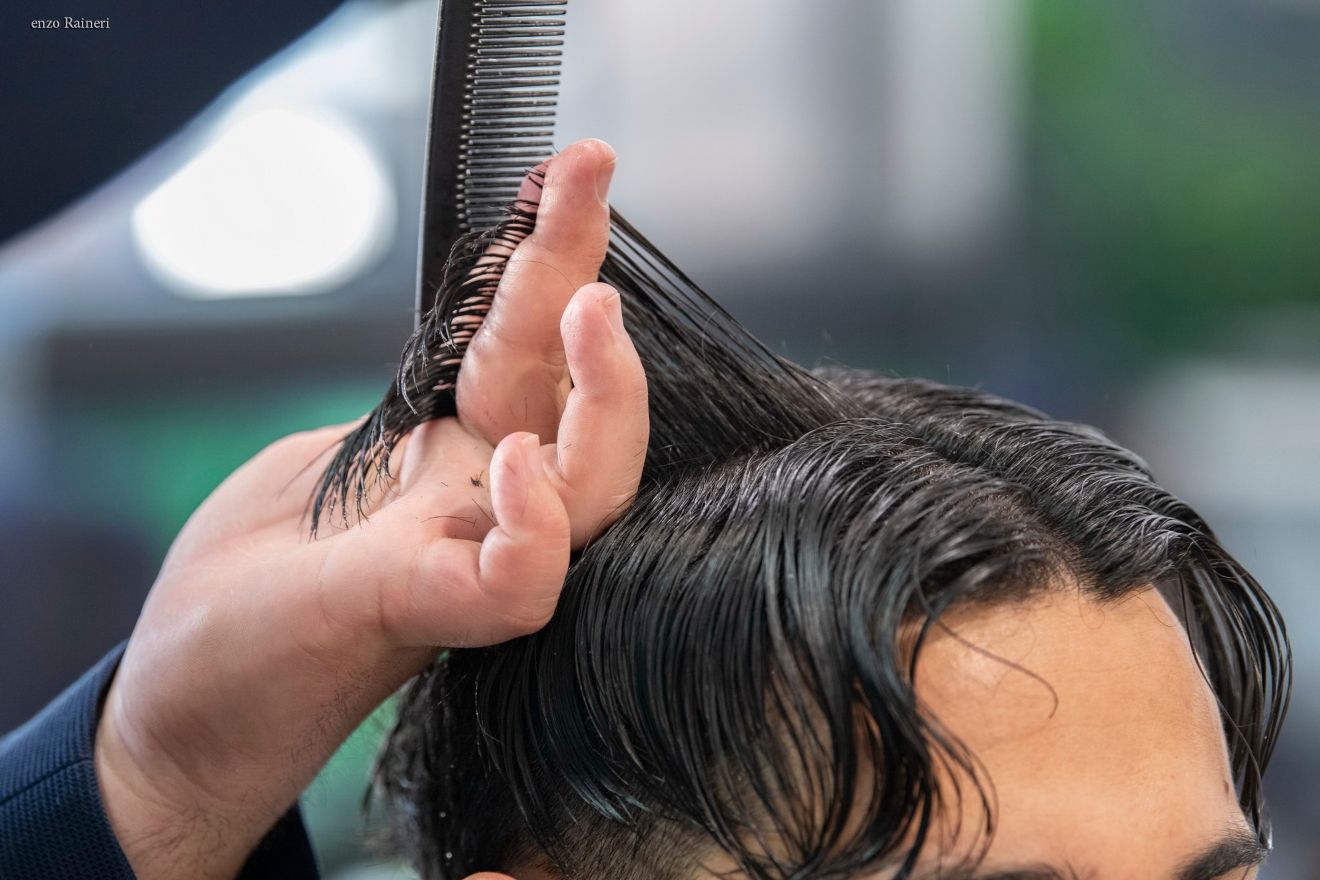Un parrucchiere a Fiumefreddo di Sicilia accorcia con perizia i capelli scuri di una cliente con precisione, usando pettine e forbici. Le mani afferrano delicatamente ciocche di capelli, sullo sfondo di uno sfondo interno delicatamente sfocato che sussurra di estrema professionalità.