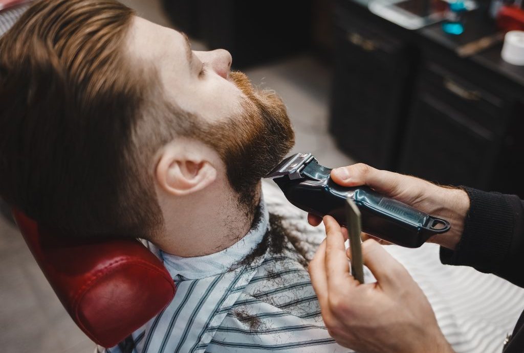 Una persona con una pettinatura ben pettinata si rilassa su una sedia da barbiere al Barbiere a Fiumefreddo di Sicilia, mentre un barbiere esperto gli rifinisce la barba con le forbici. I ritagli di capelli sono sparsi sul suo mantello a righe.