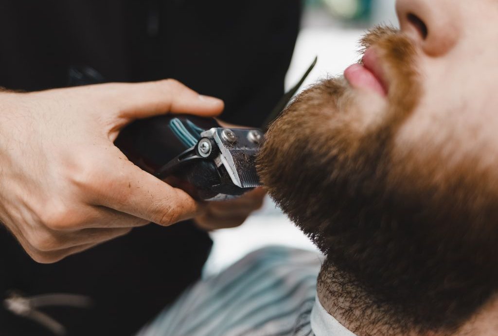 Primo piano di un Barbiere a Fiumefreddo di Sicilia che rifinisce con perizia la folta barba castana di un uomo con un tagliacapelli elettrico. L'uomo indossa una mantella da barbiere a righe e l'attenzione è rivolta alle mani che tengono il tagliacapelli e alla parte inferiore del viso, a dimostrazione di precisione e abilità.