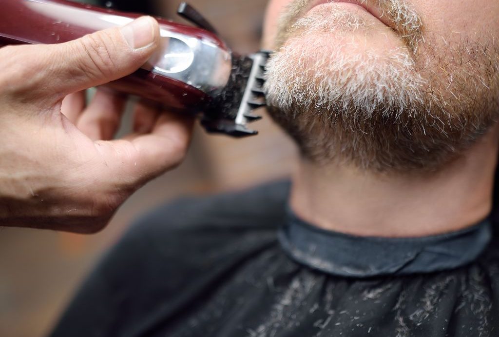 Primo piano di una persona che si fa tagliare la barba con un tagliacapelli elettrico da Barbiere a Fiumefreddo di Sicilia. L'individuo indossa una mantella da salone nera, con ritagli di capelli visibili. L'attenzione è rivolta al viso e al tagliacapelli di precisione in azione.