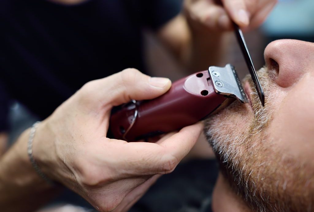 Un primo piano di una persona che riceve una rifinitura della barba da Barbiere a Fiumefreddo di Sicilia. Le mani tengono abilmente il tagliacapelli e il pettine, modellando sapientemente i baffi. I peli del viso circostanti mostrano accenni di grigio, mentre lo sfondo rimane abilmente sfocato.