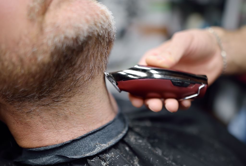 Un primo piano di un barbiere di Barbiere a Fiumefreddo di Sicilia che rifinisce la barba grigia di un uomo con un tagliacapelli elettrico. I ritagli di capelli spolverano la cappa nera del barbiere mentre la mano ferma del barbiere guida il tagliacapelli lungo il collo dell'uomo.