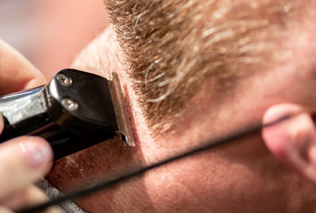 In primo piano, una persona si gode un taglio di capelli con le tosatrici elettriche eseguito dall'abile Barbiere a Fiumefreddo di Sicilia. Le tosatrici rifiniscono sapientemente i capelli sulla nuca, mostrando un taglio netto e preciso.