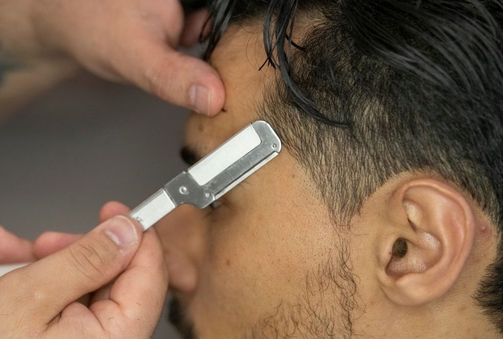 Un barbiere di Fiumefreddo di Sicilia usa abilmente un rasoio a mano libera per modellare l'attaccatura dei capelli di un cliente. Il cliente, visto di lato, sfoggia capelli scuri e una barba curata, mentre le mani del barbiere guidano delicatamente il rasoio per la precisione.