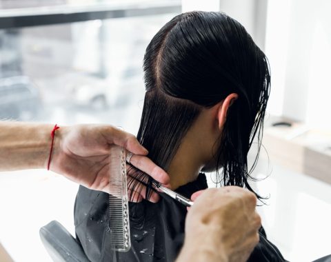 Una persona con lunghi capelli neri è da un parrucchiere a Fiumefreddo di Sicilia, per farsi tagliare i capelli. Le mani del parrucchiere maneggiano abilmente forbici e pettine, mentre la grande finestra sullo sfondo offre uno scorcio di paesaggio esterno sfocato.