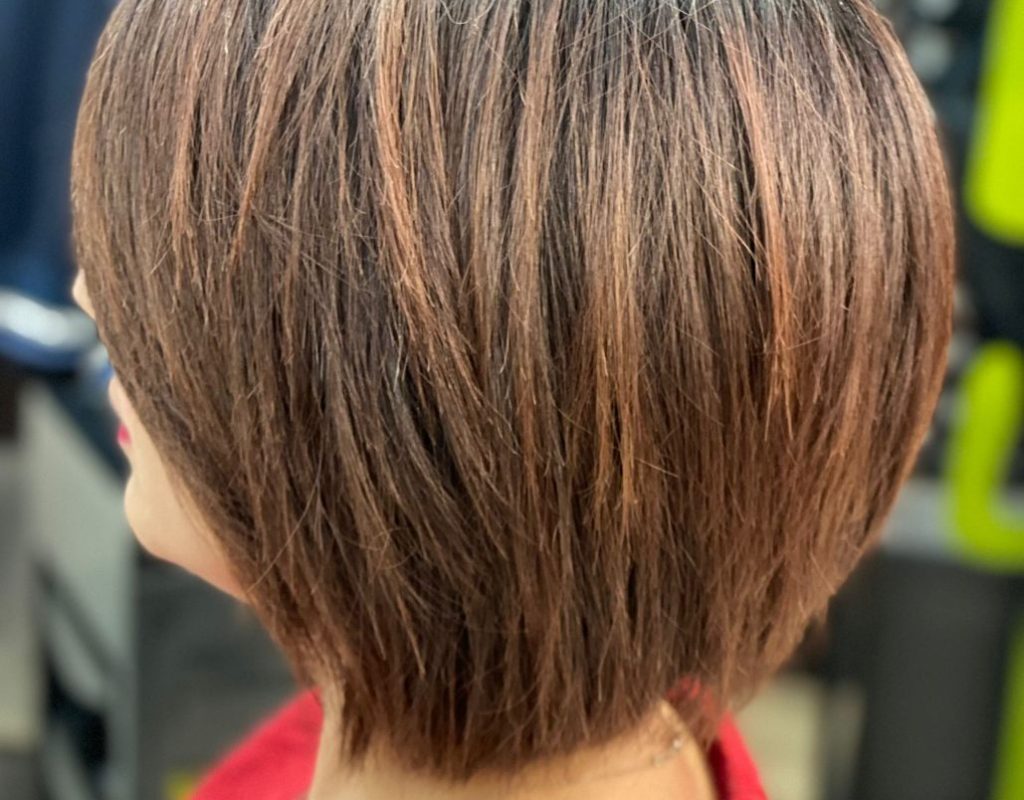 Una persona con un caschetto corto e scalato con riflessi chiari, vista da dietro. Indossa un top rosso in un ambiente interno.