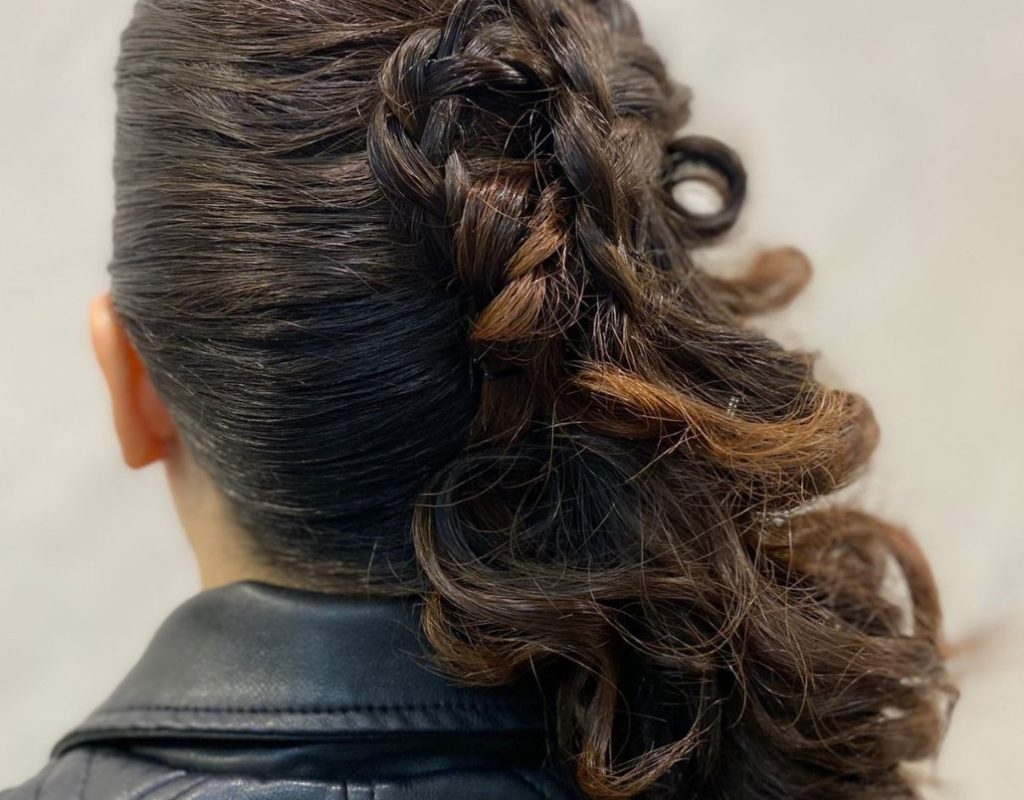 Una donna con capelli scuri e ondulati, acconciati in un elaborato updo, con trecce e riccioli, che indossa una giacca di pelle nera. La foto è scattata da dietro, e mette in mostra l'intricata acconciatura.