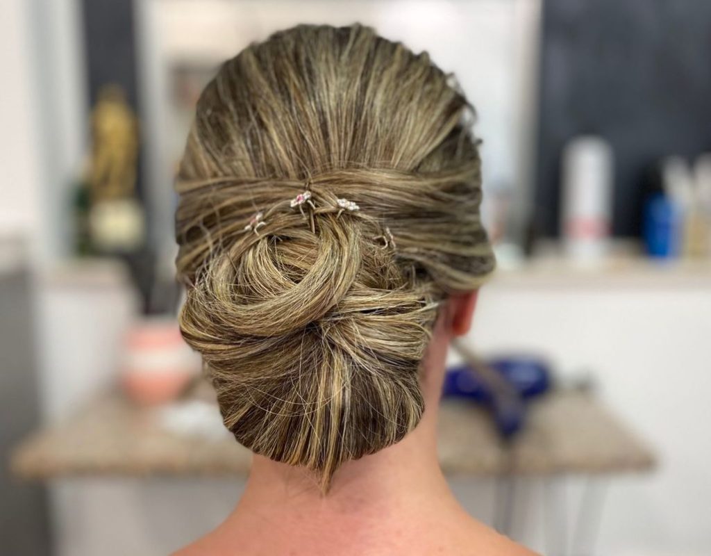 Una donna con i capelli biondi raccolti in un elegante chignon, fermati con forcine argentate, è vista da dietro. Indossa un top con fantasia blu navy e bianco. Lo sfondo è un interno di un salone delicatamente focalizzato.