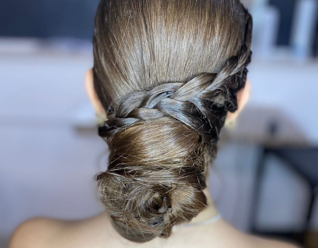 Una donna con i capelli castani lisci raccolti in un elegante chignon intrecciato. Sono visibili solo la parte posteriore della testa e le spalle, con uno sfondo sfocato che suggerisce un ambiente interno.