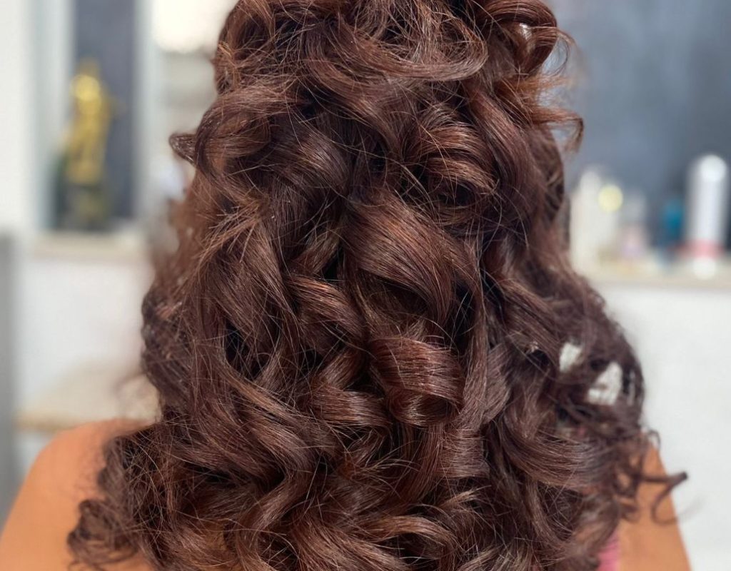 Primo piano di una persona con lunghi capelli castani ricci, acconciati in voluminosi riccioli. La persona indossa un top colorato con spalline sottili. Lo sfondo è sfocato, mostra un muro grigio e alcuni oggetti indistinti.