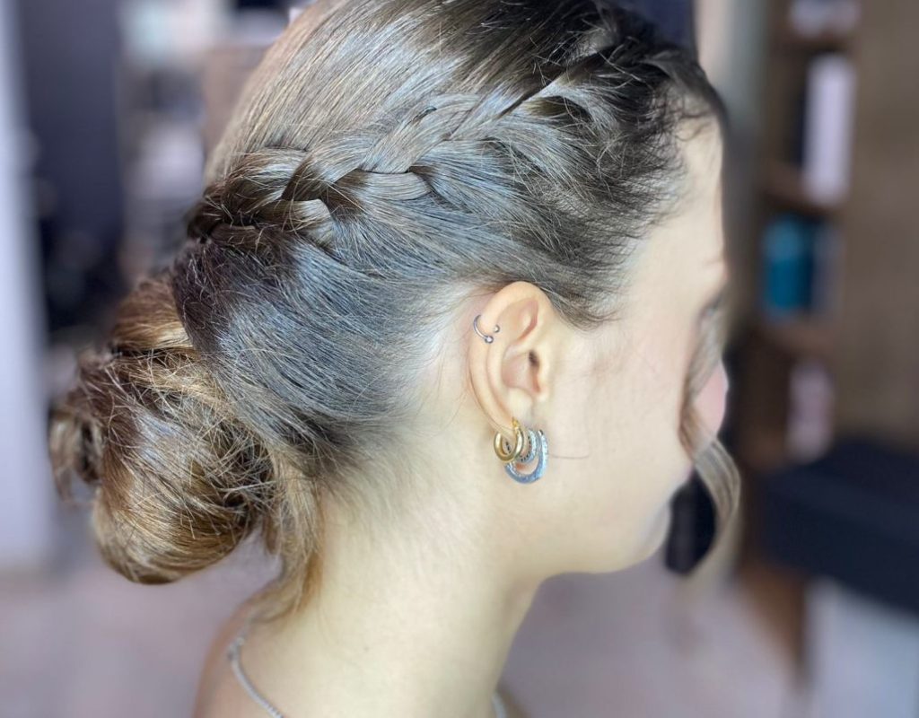 Una persona con capelli castano chiaro raccolti in una treccia è vista di lato. Indossa orecchini a cerchio e una collana sottile. Lo sfondo mostra l'interno sfocato di una stanza moderna con pareti e scaffali scuri.