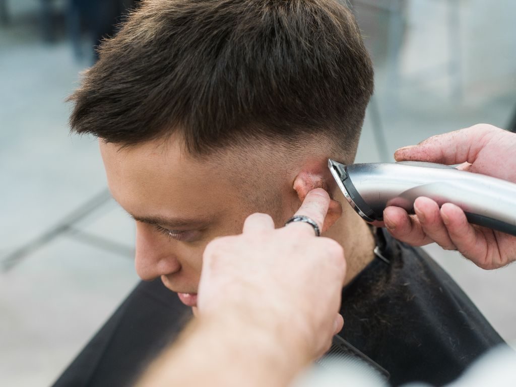 Primo piano di una persona che si fa tagliare i capelli. Un barbiere usa le macchinette per tagliare i capelli ai lati della testa. La persona ha i capelli corti e scuri e indossa un mantello nero.
