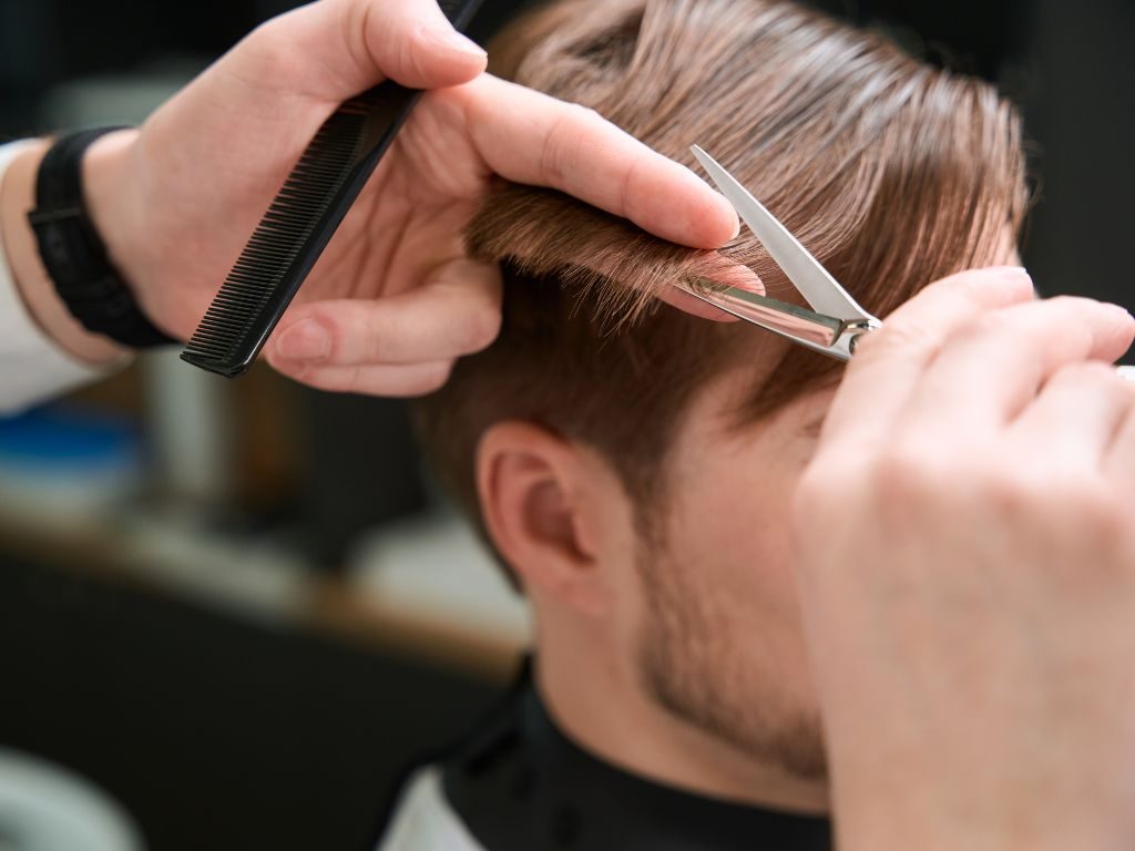 Una persona che si fa tagliare i capelli, con un primo piano di mani che usano forbici e pettine per tagliare i capelli castani. L'attenzione dello stilista è rivolta alla precisione e ai dettagli in questo ambiente da salone.