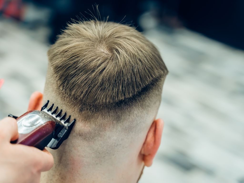 Una persona con capelli castano chiaro si sta tagliando i capelli. Il parrucchiere sta usando le macchinette per radere i lati e la parte posteriore della testa, lasciando la parte superiore più lunga. I capelli sulla parte superiore formano una linea dritta sopra le orecchie, creando un effetto taglio a scodella.