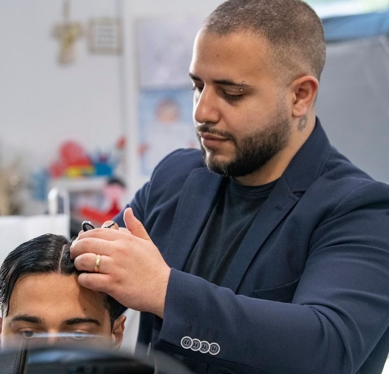 In un salone di Fiumefreddo di Sicilia, un barbiere tatuato con un anello taglia con perizia i capelli bagnati di un cliente. Il cliente siede parzialmente visibile sulla sedia, circondato da attrezzature e decorazioni del salone che contribuiscono a creare un'atmosfera accogliente.