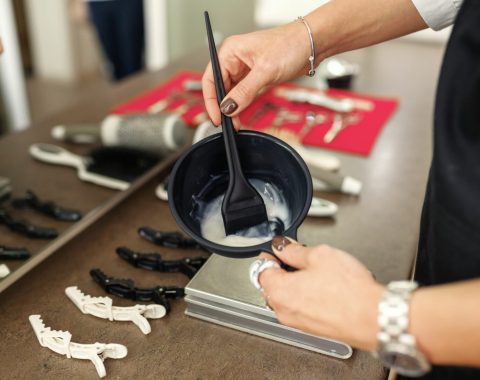 Una persona è in piedi come un parrucchiere a Fiumefreddo di Sicilia, con un pennello sopra una ciotola di tintura cremosa per capelli, pronta per essere mescolata. Gli strumenti per lo styling dei capelli e le mollette sono disposti ordinatamente sul tavolo. L'individuo indossa un orologio e un braccialetto, il suo riflesso è visibile nello specchio.