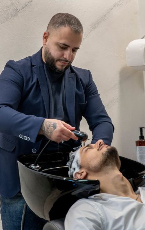 Un parrucchiere a Fiumefreddo di Sicilia, con barba e tatuaggi sulla mano sinistra, sta lavando i capelli a una cliente. La cliente è reclinata sulla sedia, con la testa appoggiata su un lavandino nero, e indossa una camicia chiara. Sullo sfondo, una serie di flaconi di shampoo aggiunge fascino al salone.