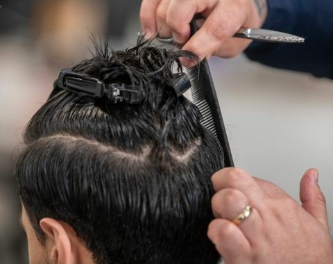 Un parrucchiere a Fiumefreddo di Sicilia usa sapientemente pettine e forbici per tagliare i capelli bagnati e scuri. Le sezioni sono tenute in posizione con delle mollette, concentrandosi sulle mani esperte e sugli strumenti in azione.