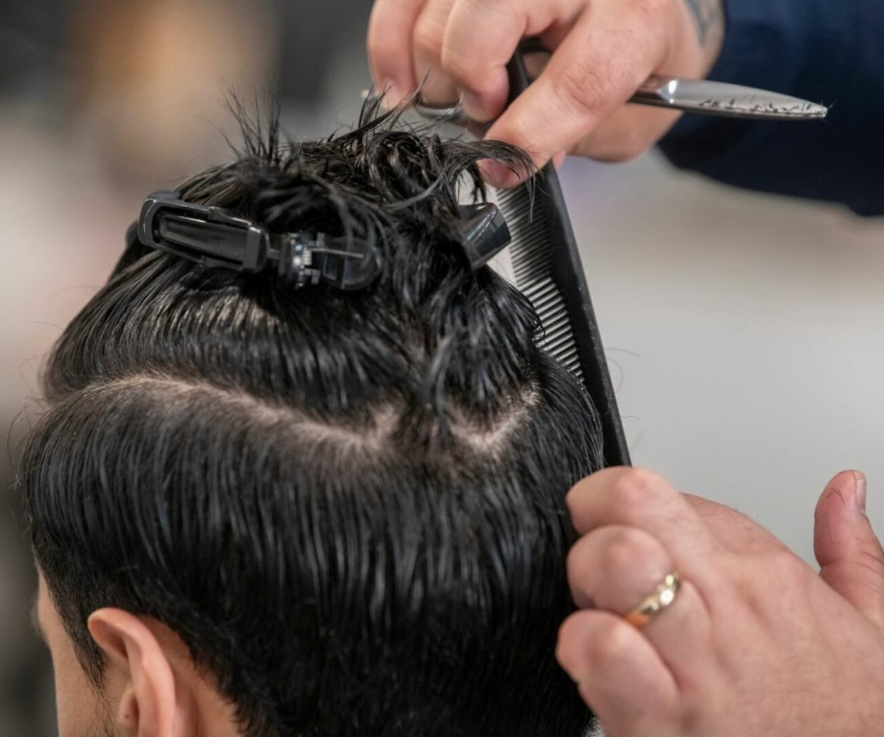 Un parrucchiere a Fiumefreddo di Sicilia usa sapientemente pettine e forbici per tagliare i capelli bagnati e scuri. Le sezioni sono tenute in posizione con delle mollette, concentrandosi sulle mani esperte e sugli strumenti in azione.